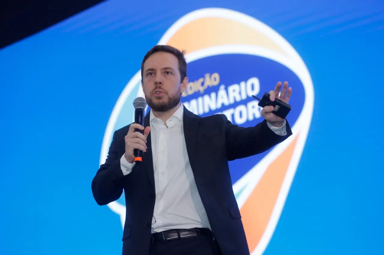 Homem de terno escuro e camisa branca faz uma apresentação em um palco, segurando um microfone em uma das mãos e um controle remoto na outra. Ao fundo, há uma tela azul com um logotipo circular desfocado relacionado a um seminário do setor. O palestrante aparece em destaque, gesticulando durante sua fala.