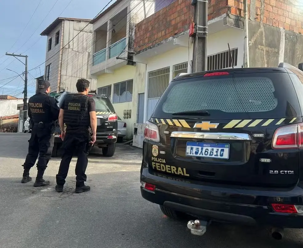 Viatura da Polícia Federal e agentes em frente a um prédio residencial, com carro oficial Chevrolet estacionado na rua durante operação, no Brasil.
