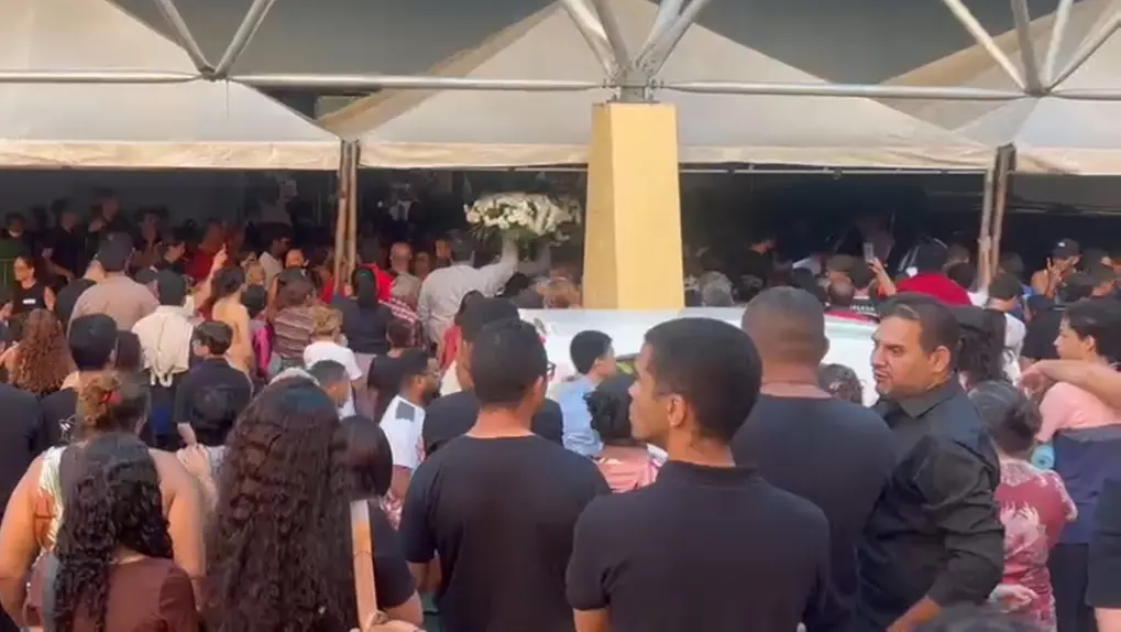 Uma grande multidão de pessoas reunida sob a estrutura de tendas brancas em uma área externa. Muitas pessoas estão de costas, olhando em direção ao centro, onde se destaca uma grande coroa de flores brancas sendo erguida acima das cabeças.