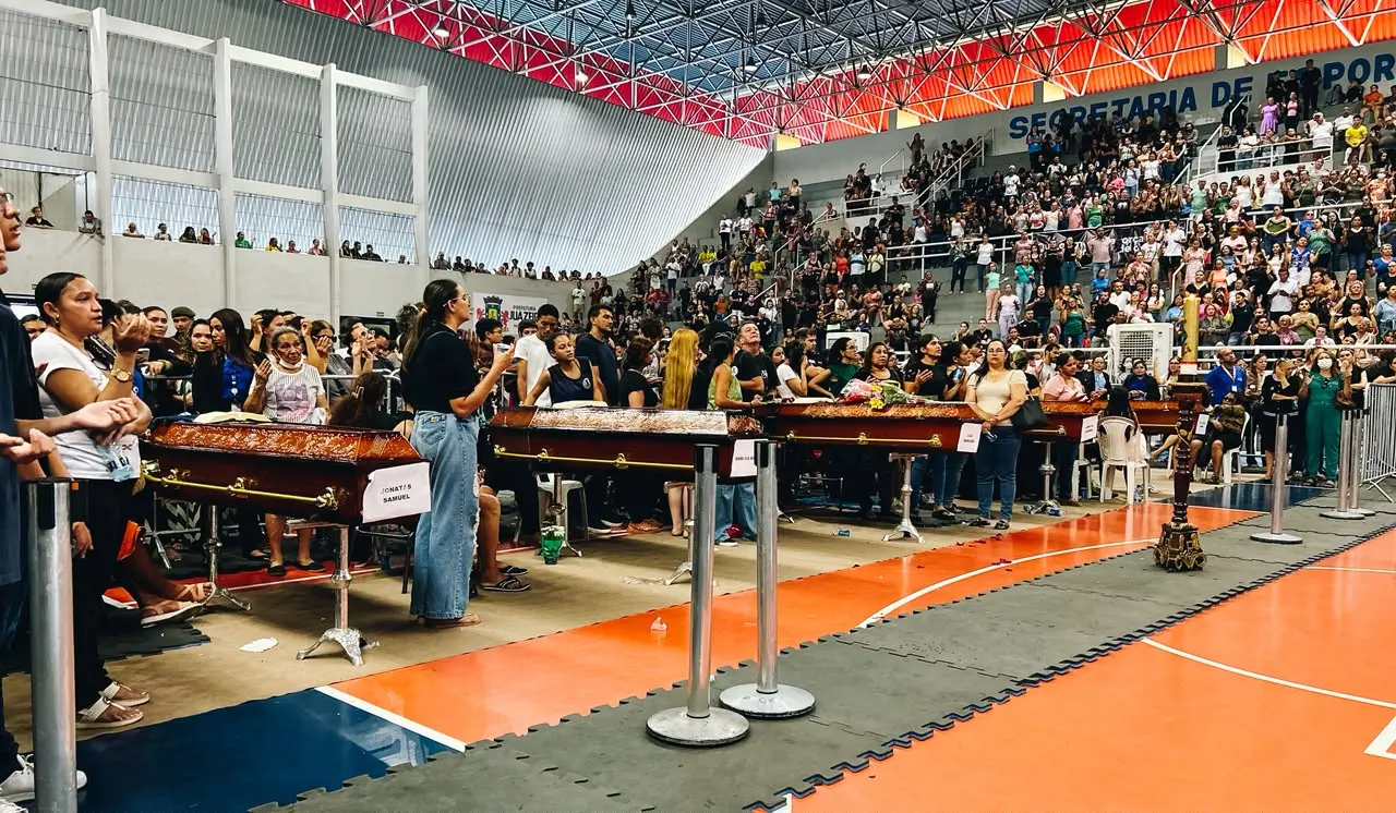 Foto mostra multidão de pessoas na arquibancada de um ginásio esportivo. Na quadra, caixões são enfileirados com os corpos de vítimas de acidente. Pessoas rezam no entorno dos caixões.
