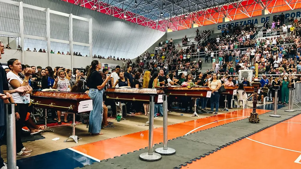 Foto mostra multidão de pessoas na arquibancada de um ginásio esportivo. Na quadra, caixões são enfileirados com os corpos de vítimas de acidente. Pessoas rezam no entorno dos caixões.