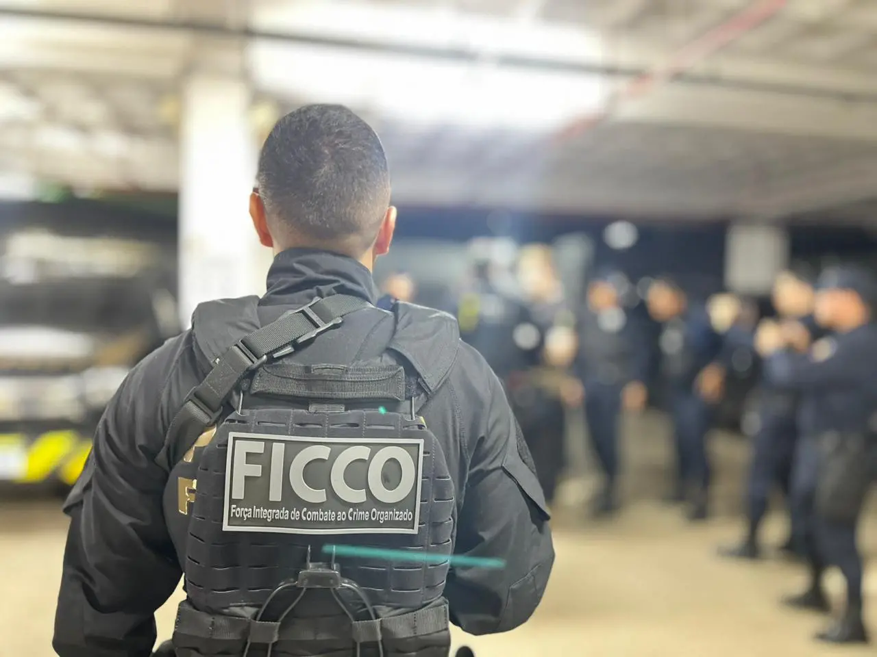 Pessoa vista de costas usando colete tático preto com identificação “FICCO – Força Integrada de Combate ao Crime Organizado”, em ambiente interno com outras pessoas ao fundo desfocadas.