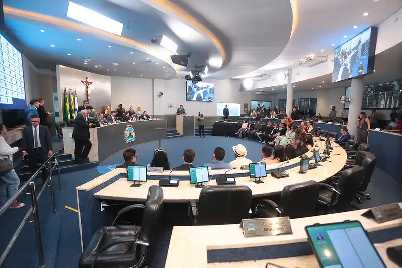 Plenário da Câmara, com mesa redonda de madeira e diversas pessoas à frente.