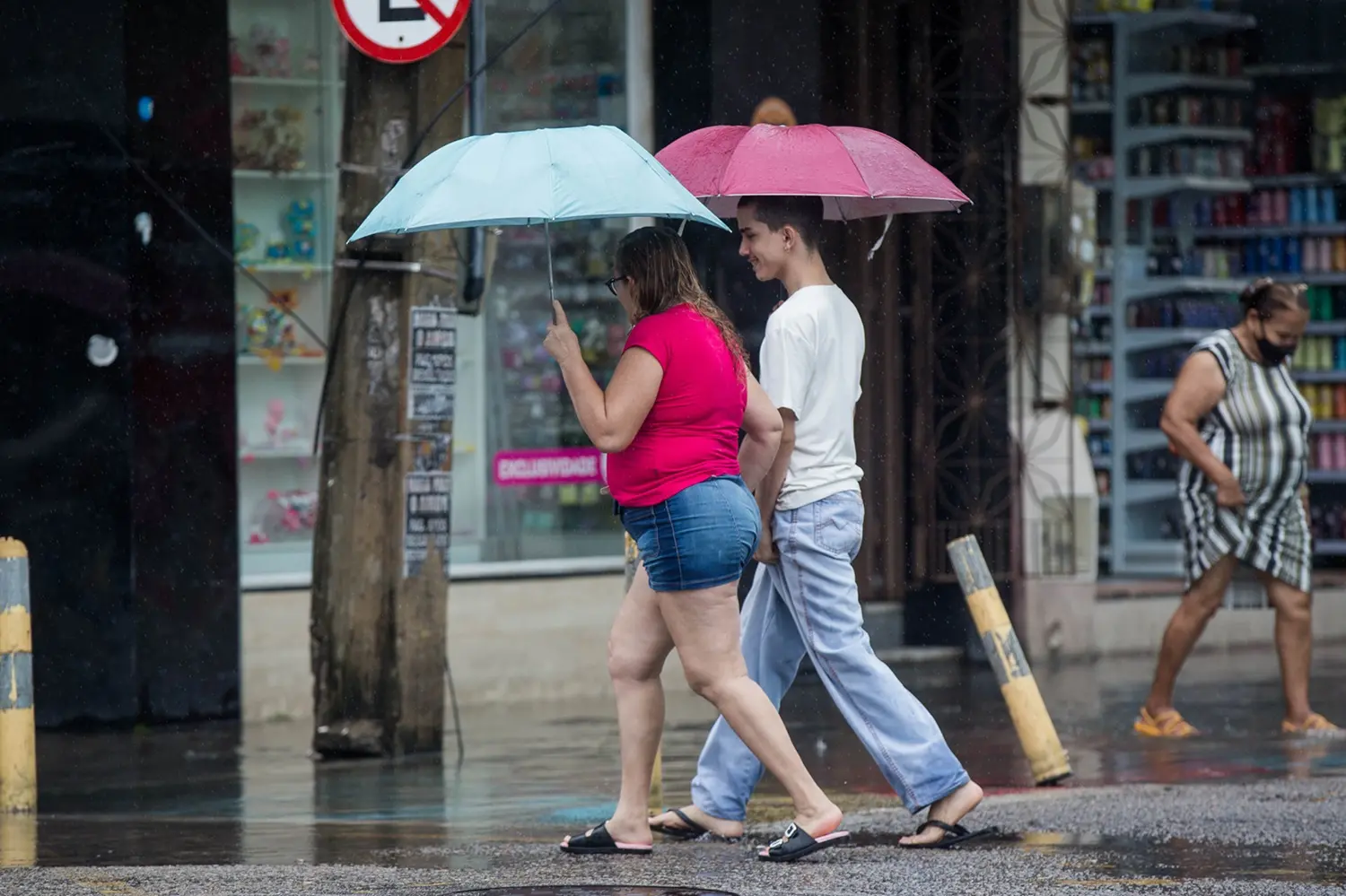 Pedestres caminhando sob chuva em uma calçada urbana molhada no Centro de Fortaleza. Em destaque, um casal anda de perfil: uma mulher com blusa rosa e shorts jeans segura um guarda-chuva azul claro, enquanto um jovem de camiseta branca e calça jeans segura um guarda-chuva vermelho escuro.