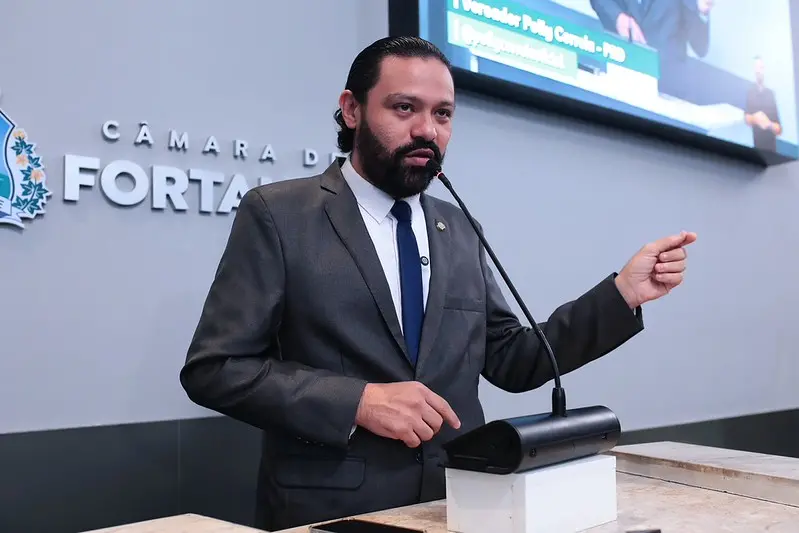 Homem branco de cabelos longos pretos, barba e bigode pretos, usando terno cinza e discursando em tribuna.