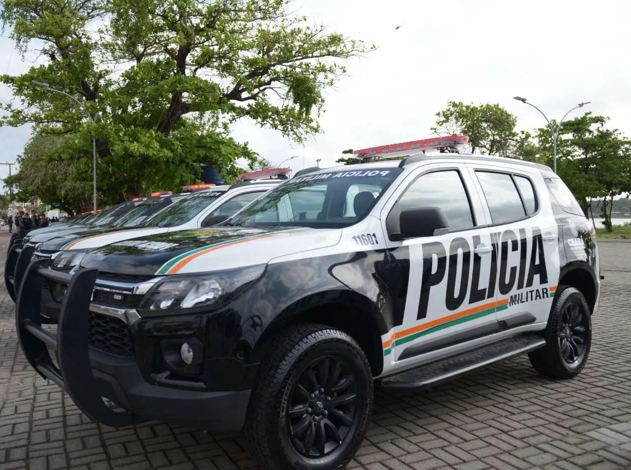 Viaturas da Polícia Militar do Ceará estacionadas lado a lado em área pavimentada, com luzes no topo, árvores ao fundo e céu nublado.