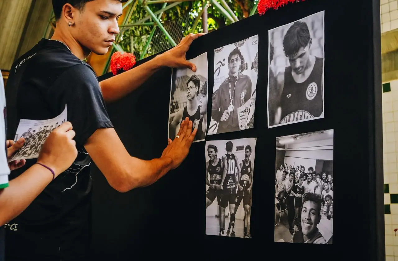 Um jovem de camiseta preta fixa fotografias em preto e branco em um painel expositor escuro. As imagens coladas retratam estudantes em competições esportivas e momentos em sala de aula. Outra pessoa aparece parcialmente à esquerda segurando um folheto impresso.