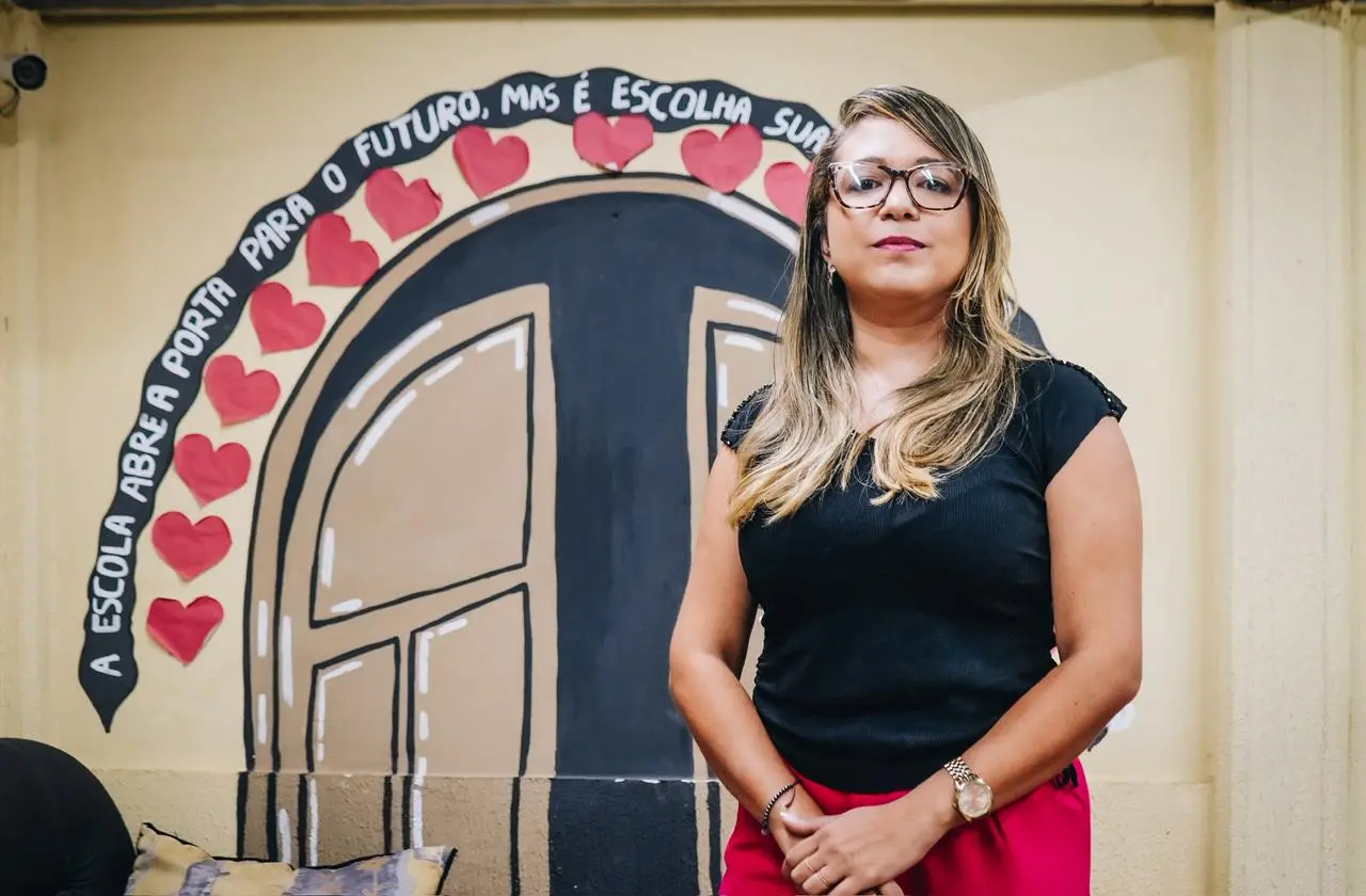 Uma mulher de óculos, cabelos longos e blusa preta posa em frente a um mural escolar colorido. O fundo pintado na parede simula um portal preto adornado com corações vermelhos e uma frase arqueada. A frase em destaque diz: A ESCOLA ABRE A PORTA PARA O FUTURO, MAS É ESCOLHA SUA.