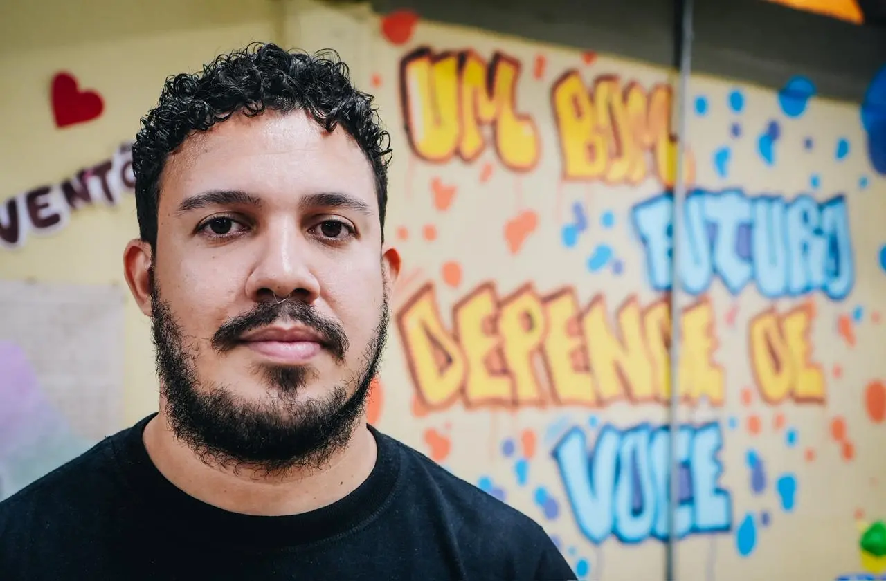 Retrato em plano fechado de um homem jovem com cabelos cacheados pretos, barba e um piercing no septo. Ele veste uma camiseta preta e olha diretamente para a câmera com uma expressão serena. Ao fundo, há uma parede amarela decorada com grafites coloridos em tons de azul e laranja.