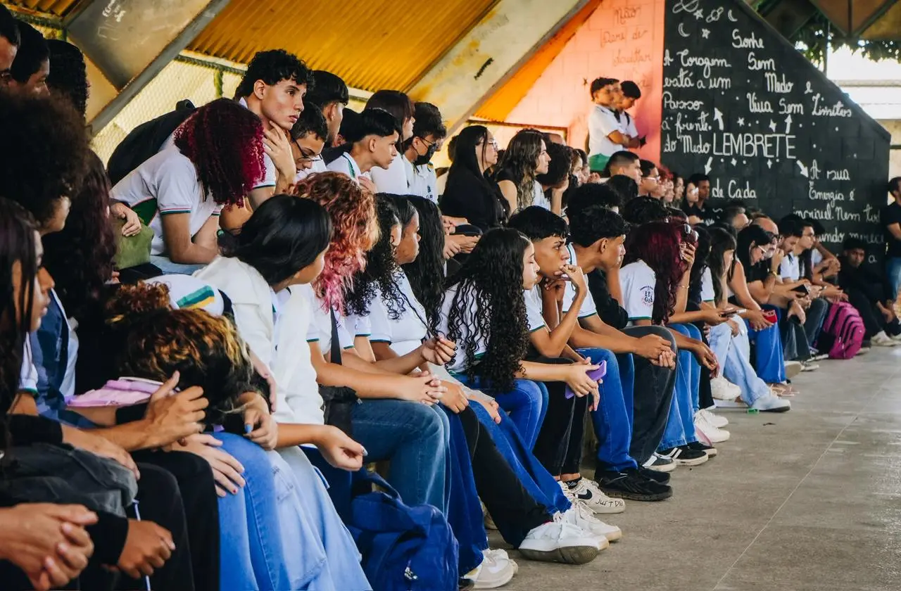 Vários jovens estão sentados em uma arquibancada coberta de cimento durante um evento escolar. A maioria veste uniformes brancos e calças jeans, olhando atentamente em uma mesma direção. No fundo, uma parede preta exibe frases motivacionais escritas à mão com giz branco.