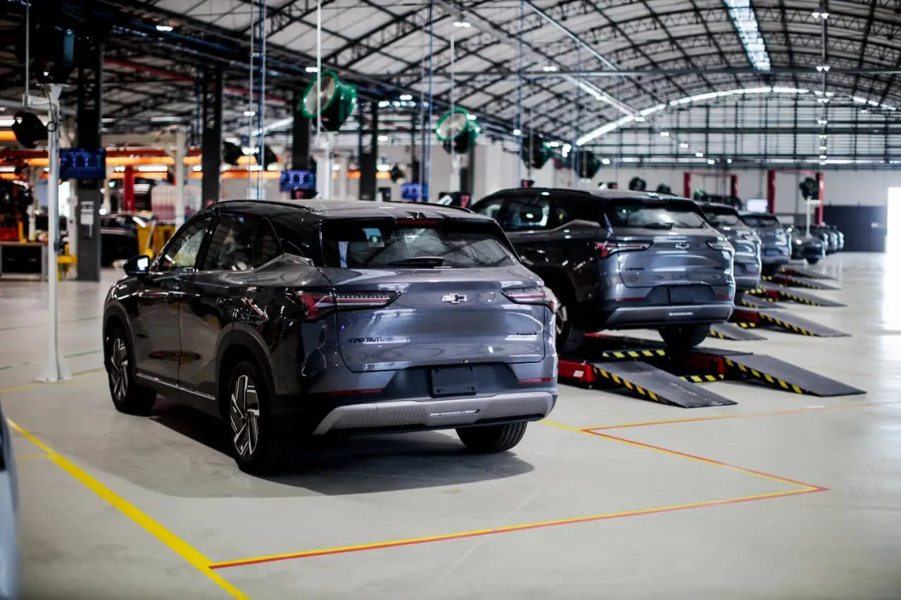 SUVs elétricos alinhados em uma área de inspeção e testes dentro de uma fábrica automotiva. Os veículos estão distribuídos em estações de trabalho sob um galpão amplo, com equipamentos de suporte e sinalização industrial ao redor.