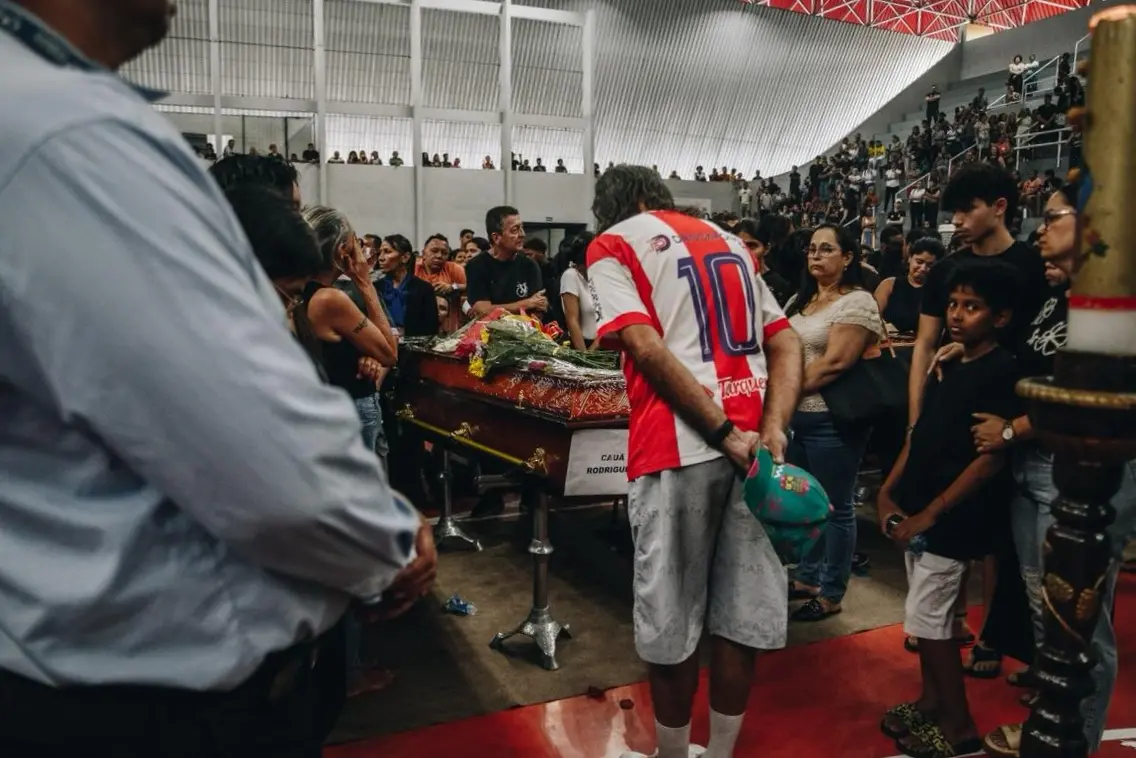 Pessoas se reúnem em um ginásio para prestar homenagens diante de um caixão, com destaque para um homem de camisa esportiva de costas.