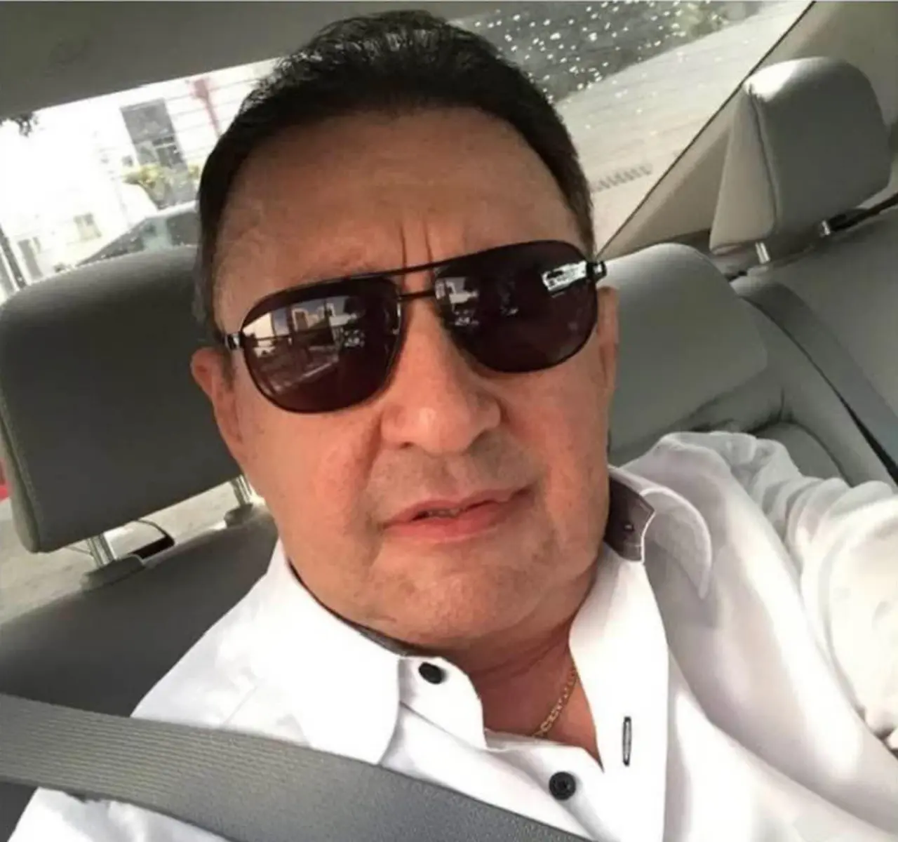 Homem branco de cabelos pretos, usando óculos escuros e blusa branca, sentado em banco de carro.