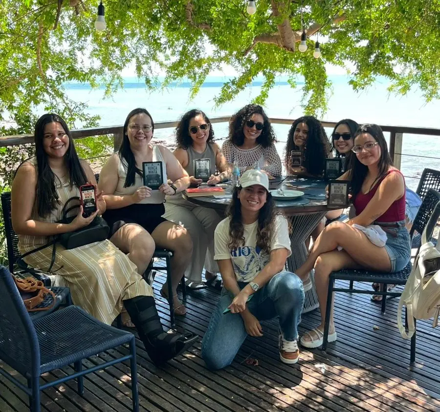 Grupo de mulheres participa de um encontro de clube de leitura em um deck com vista para o mar. Sentadas ao redor de uma mesa ao ar livre, elas exibem exemplares de livros enquanto posam para a foto sob a sombra de árvores, em ambiente descontraído e ensolarado.