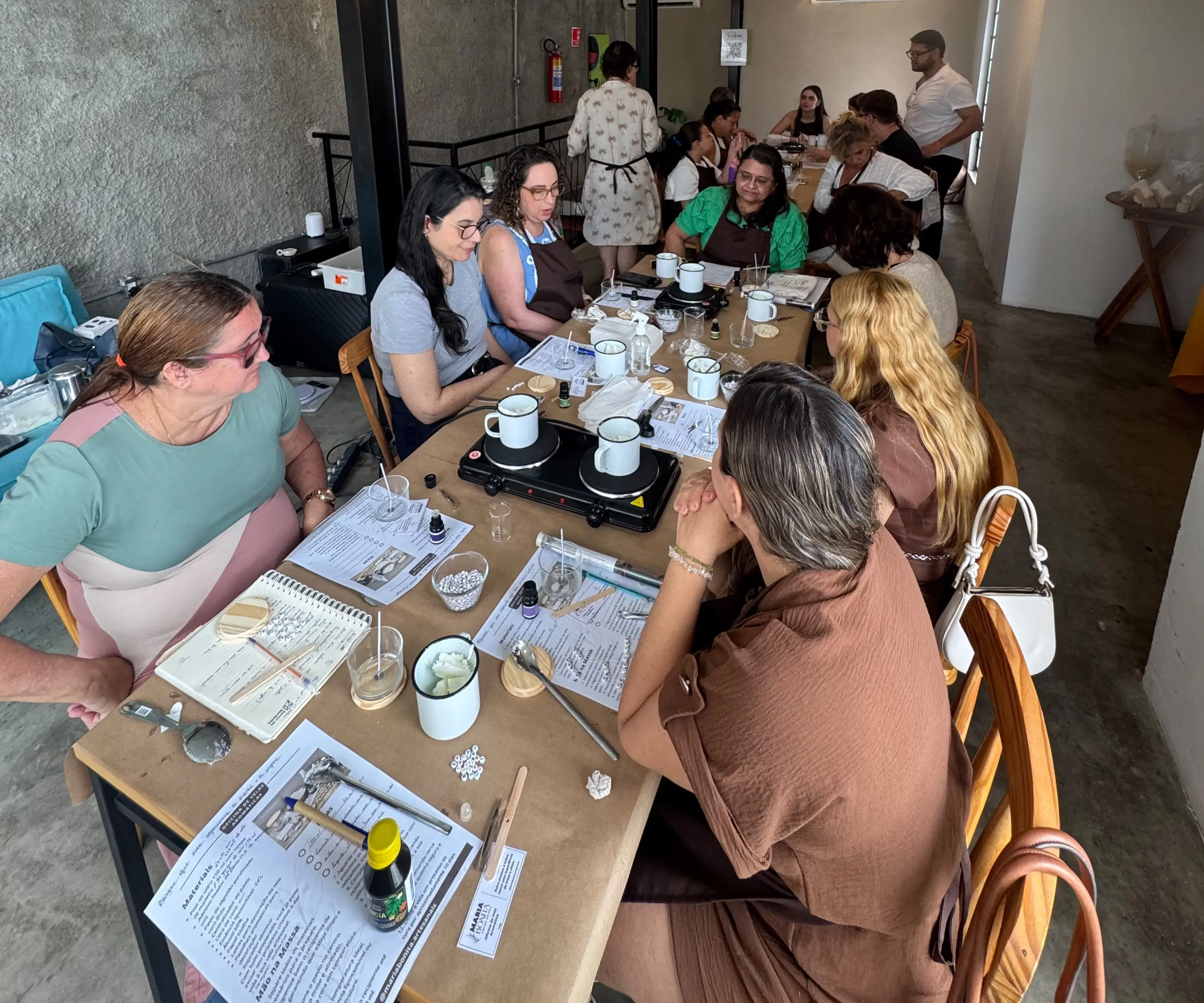 Grupo de participantes acompanha uma atividade em uma cafeteria, reunido em torno de uma mesa com materiais de trabalho, cadernos e recipientes aquecidos. O encontro ocorre em ambiente acolhedor e favorece a interação entre os participantes por meio de uma experiência coletiva.