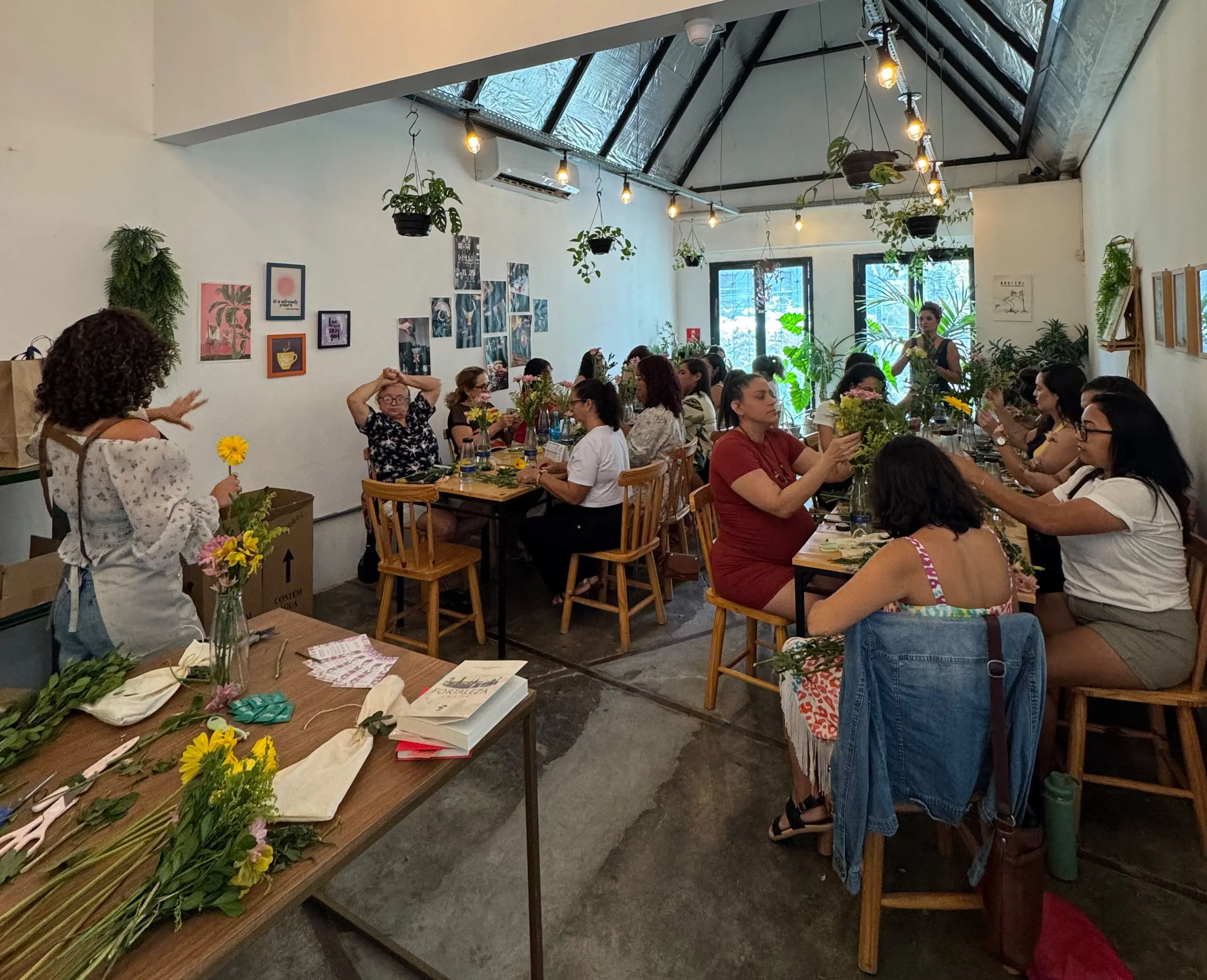 Pessoas participam de uma oficina de arranjos florais em uma cafeteria. Sentadas em mesas coletivas, elas manuseiam flores e folhagens enquanto recebem orientações da facilitadora, em um espaço iluminado e decorado com plantas.