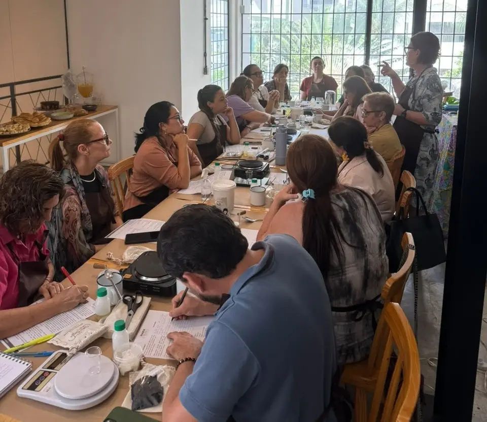Participantes de um clube de leitura ocupam uma grande mesa em uma cafeteria durante uma discussão literária. Enquanto algumas pessoas fazem anotações, outras acompanham a fala de uma mediadora em pé, em um ambiente de troca de ideias e convivência cultural.