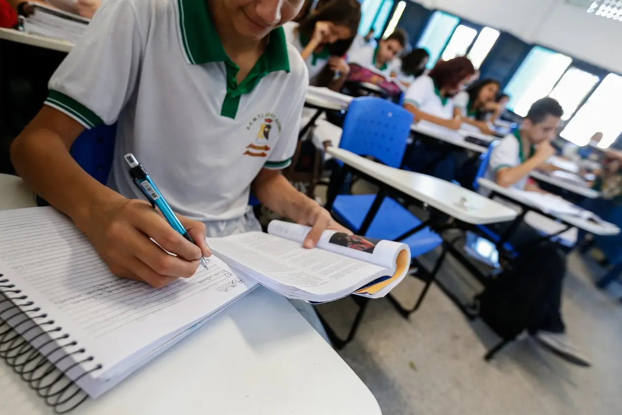 Estudante em primeiro plano escreve em um caderno espiral com uma caneta azul enquanto consulta um livro aberto. Ele usa uma camiseta de uniforme escolar branca com gola verde e um logotipo bordado no peito. Ao fundo, outros alunos estão sentados em carteiras azuis individuais, concentrados em suas atividades na sala de aula.