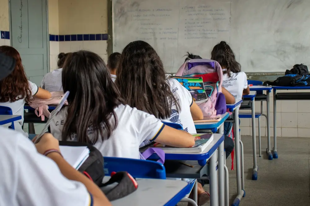 Sala de aula com estudantes sentados em carteiras individuais, vistos de trás, escrevendo e olhando para um quadro branco com anotações ao fundo; mochilas e cadernos estão sobre as mesas.