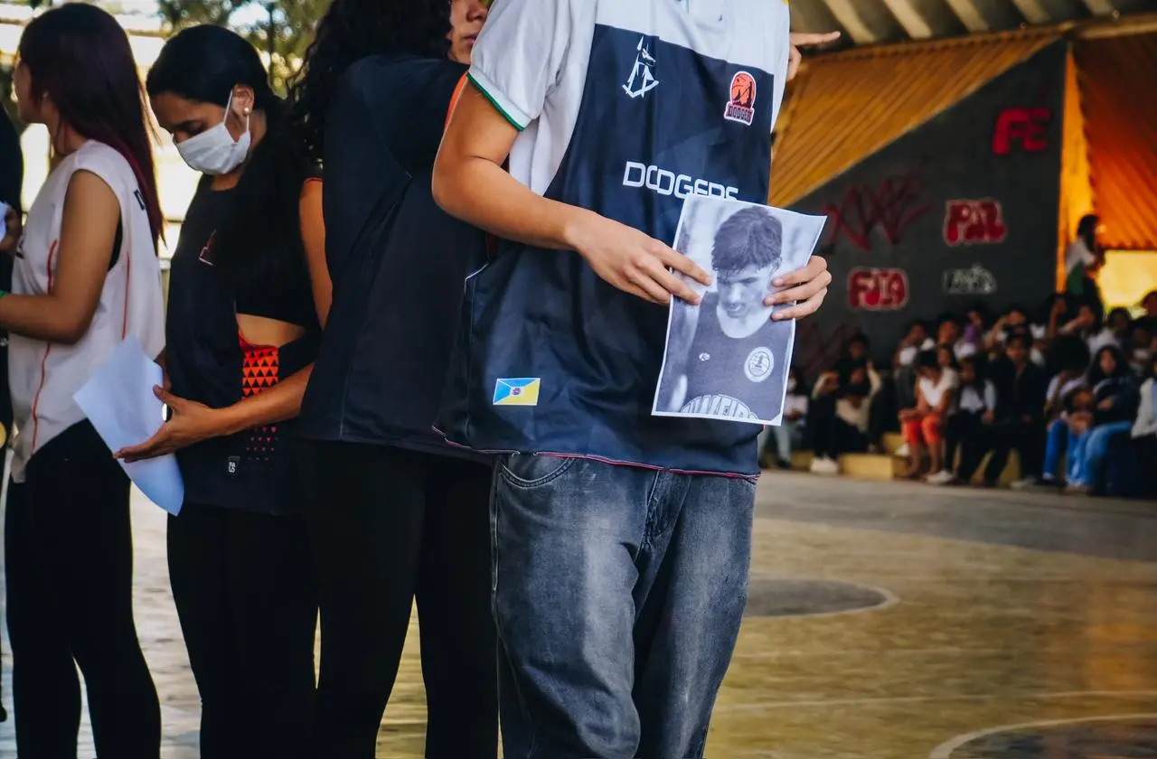 Na imagem, fotografia em plano médio de pessoas em pé em uma quadra poliesportiva coberta. No centro, em primeiro plano, uma pessoa veste uma camiseta azul-escura com a palavra 
