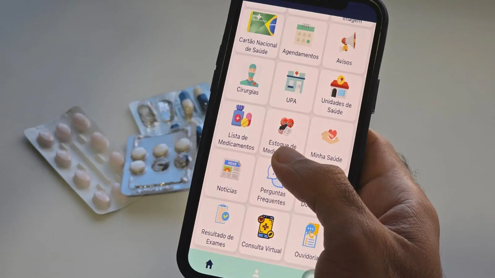 Foto de detalhe de uma mão clicando na tela de um celular mostrando um aplicativo com várias funções da área de saúde, como consultas, cirurgias, UPA e lista de medicamentos. Ao fundo, há diversas cartelas de remédios sobre uma bancada branca.