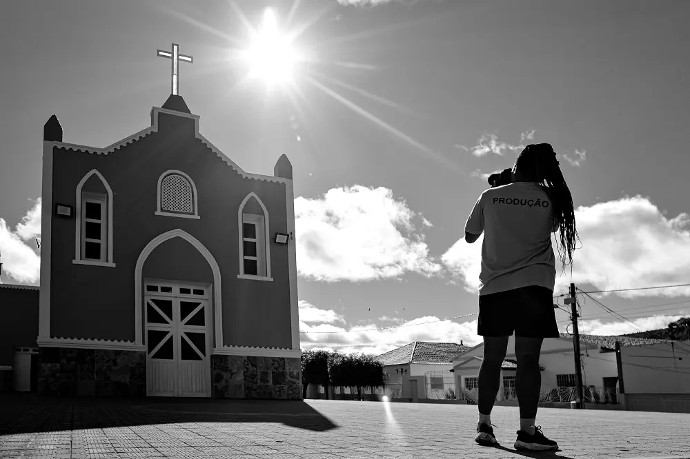 Foto em preto e branco contra a luz. Uma pessoa de costas com camiseta escrito 'produção' fotografa uma pequena igreja com uma cruz no topo. O sol brilha intensamente no céu acima da igreja.