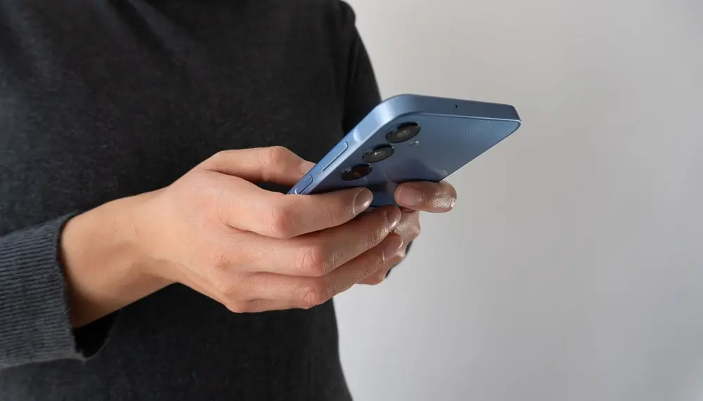 Na imagem, foto em plano fechado mostrando as mãos de uma pessoa segurando um smartphone moderno de cor azul-clara. A pessoa veste uma blusa cinza-escura de manga comprida e está posicionada de perfil, contra um fundo cinza-claro e neutro. Seus polegares estão posicionados sobre a tela, indicando que ela está digitando ou navegando no aparelho. A parte traseira do celular está visível, destacando as lentes da câmera alinhadas verticalmente.