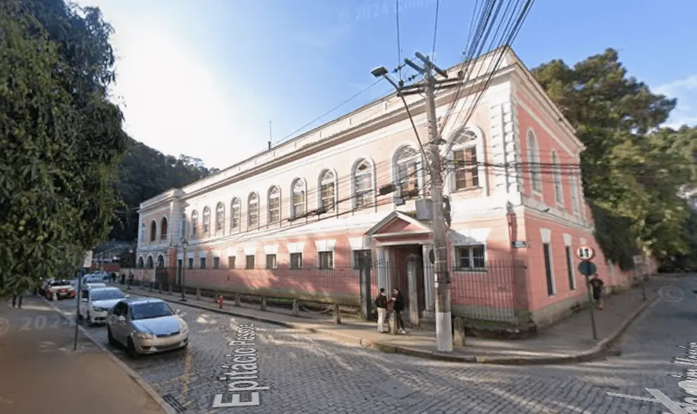 Vista externa de um edifício histórico de dois andares, pintado em tons de rosa e branco, localizado em uma esquina de rua de paralelepípedos. A construção possui diversas janelas em arco na fachada e uma entrada central com pequeno pórtico. Há carros estacionados ao longo da via, postes e fiação elétrica em frente ao prédio, além de árvores e área de vegetação ao fundo. A imagem mostra o Palácio do Grão-Pará, em Petrópolis (RJ).