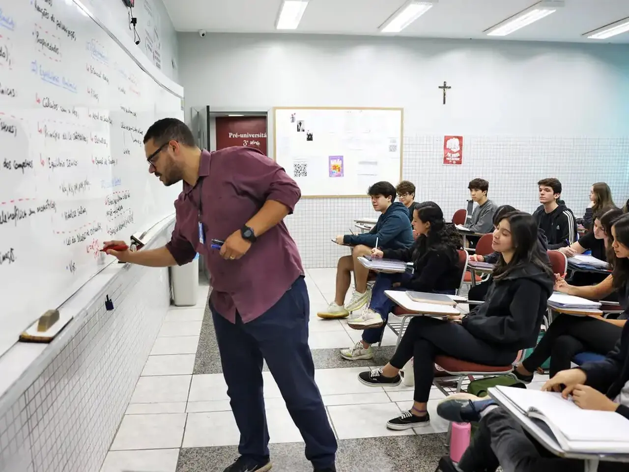 Professor pardo e de cabelo curto, usando blusa de botão roxa e calça jeans, escrevendo em quadro branco dentro de sala de aula com alunos adolescentes.