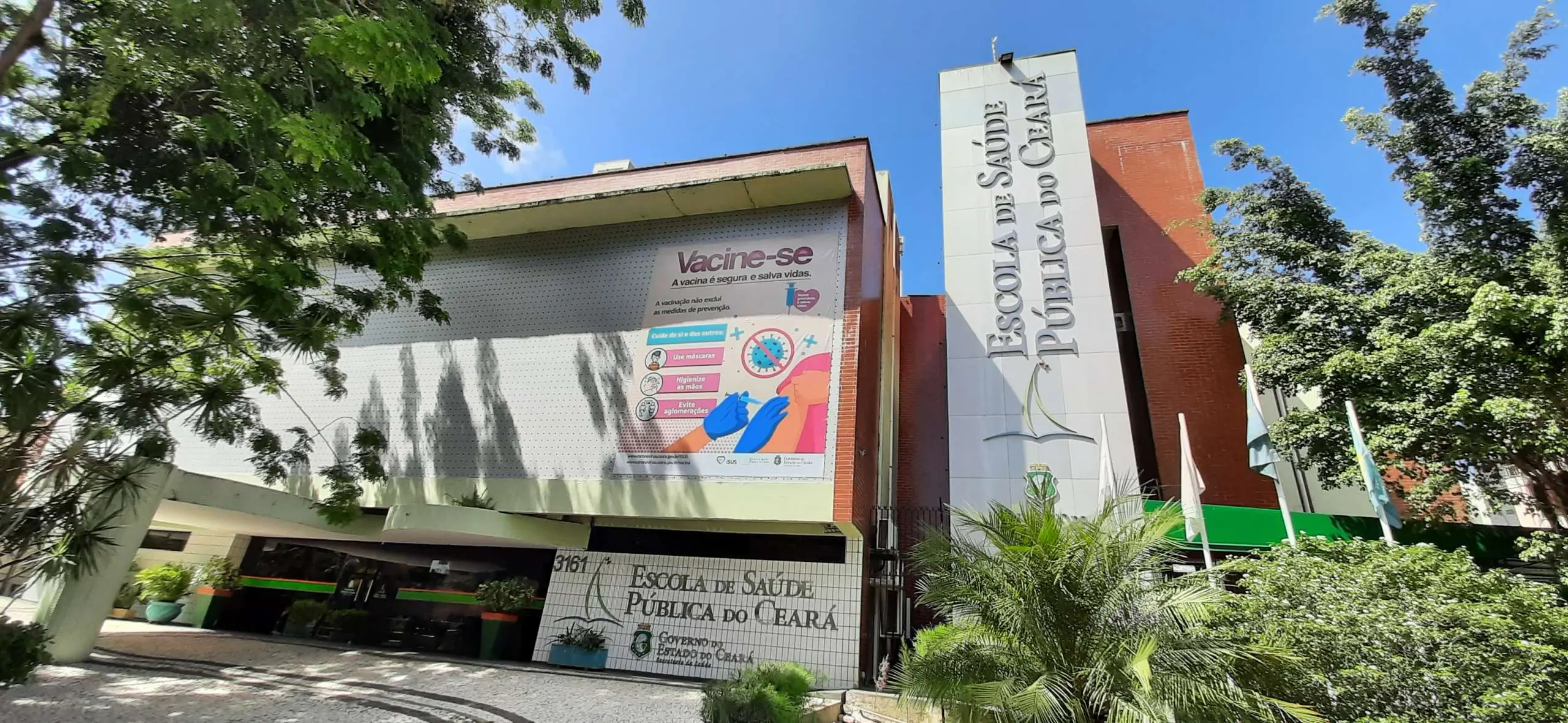 Fachada da Escola de Saúde Pública do Ceará, em Fortaleza, vista em plano aberto. O prédio possui estrutura em tons de branco e vermelho, cercada por árvores e vegetação. Na lateral, há uma torre vertical com a inscrição 