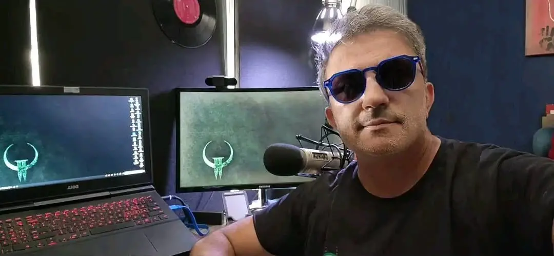 Selfie em plano médio do dublador brasileiro Figueira Júnior em um estúdio. Ele é um homem de pele clara, cabelos curtos grisalhos e barba por fazer, vestindo uma camiseta preta e óculos de sol com armação azul-escura. Ao fundo, à esquerda, há um notebook com teclado retroiluminado em vermelho e um monitor de computador ao lado, ambos exibindo um símbolo verde da franquia Quake na tela. Um microfone profissional de estúdio está posicionado à sua frente.