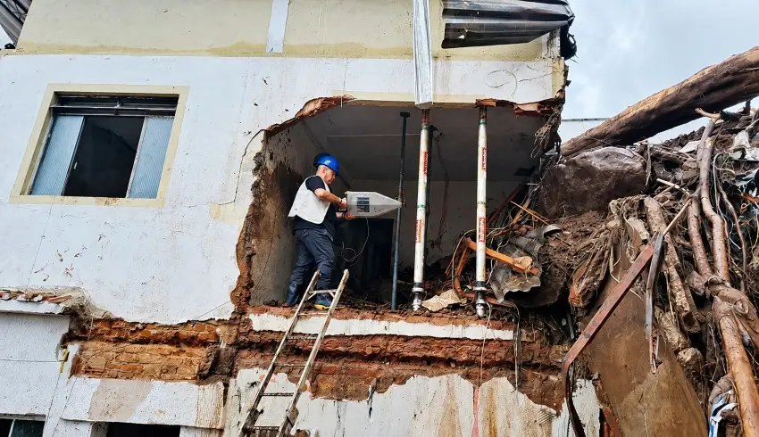 Trabalhador da construção civil com capacete e uniforme faz inspeção ou instalação elétrica em um prédio demolido, com paredes parcialmente removidas, fiação exposta e escada de acesso sob céu nublado.