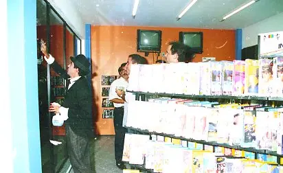 Interior da loja King Video durante a inauguração, em 1997. Pessoas observam prateleiras com fitas de vídeo e produtos em um ambiente comercial com balcão e equipamentos ao fundo.