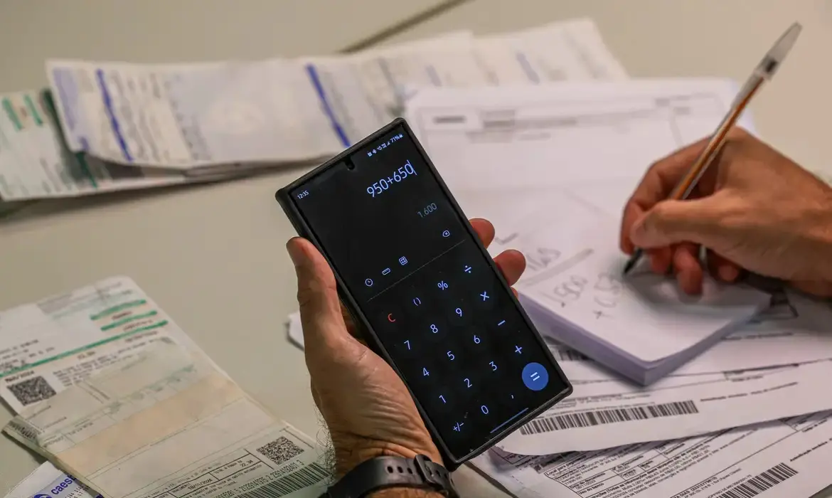 Uma pessoa segura um smartphone com o aplicativo de calculadora aberto mostrando a soma '950+650', enquanto sua outra mão faz anotações com uma caneta em um bloco de papel ao fundo. A cena é composta por diversas contas e boletos bancários espalhados sobre a mesa, criando uma atmosfera focada em planejamento financeiro e organização de despesas domésticas.