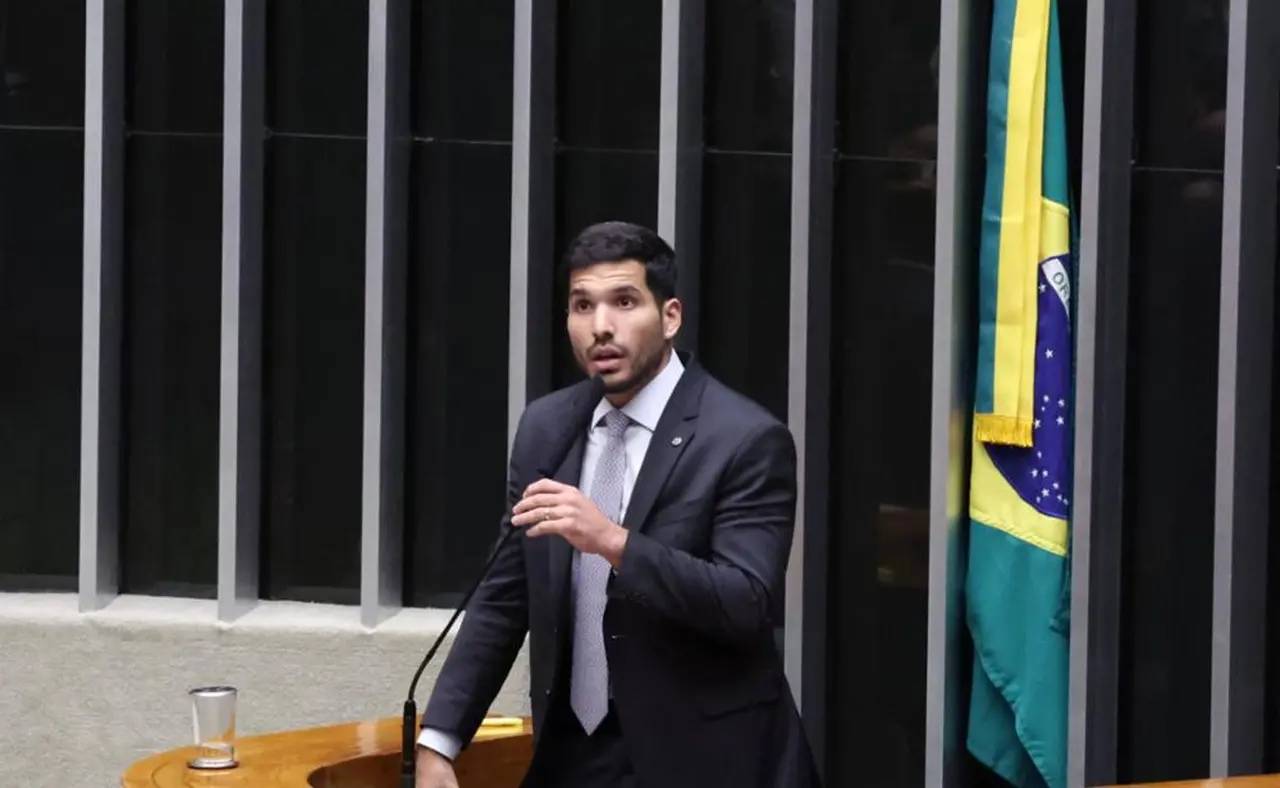 Homem branco de cabelos pretos, barba e terno discursando em palanque de plenário.