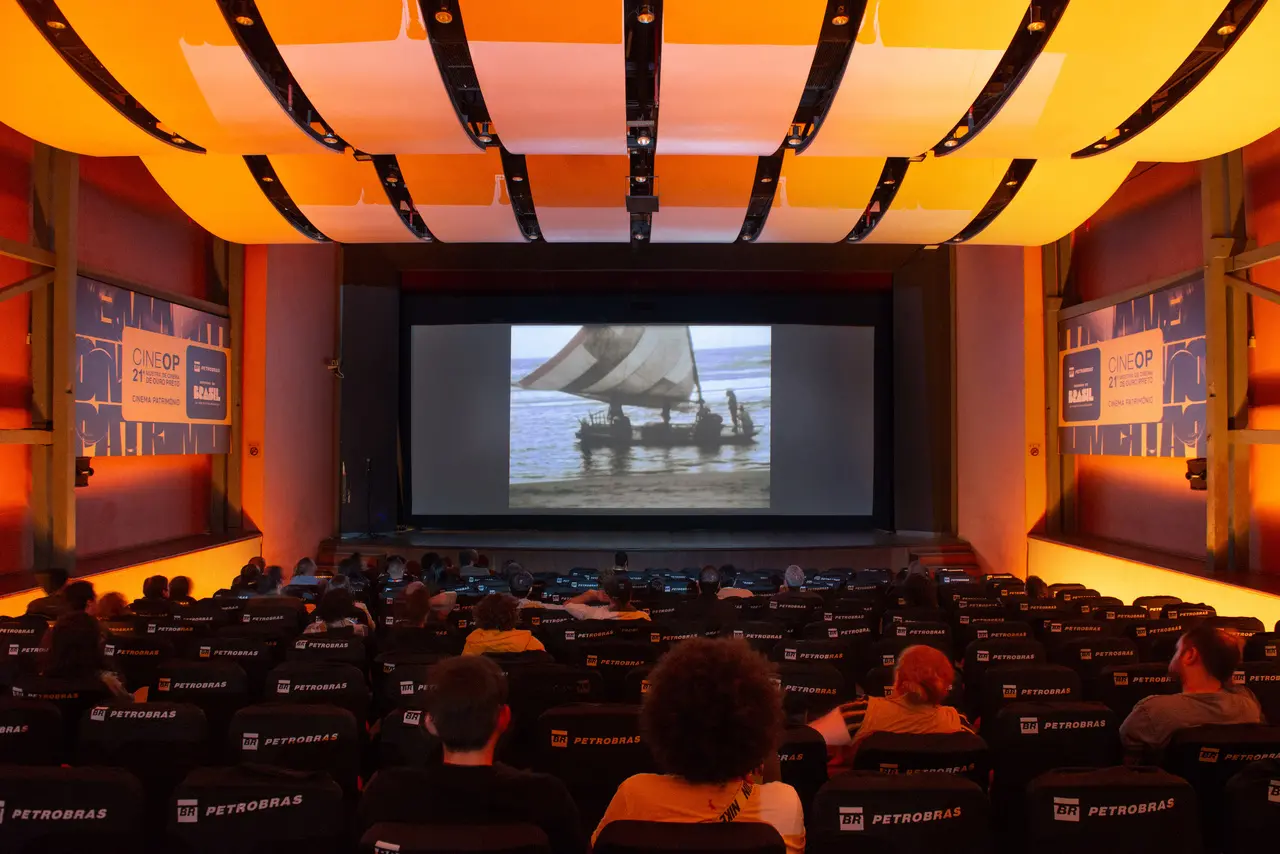 Uma plateia assiste a uma sessão de cinema dentro de um auditório iluminado por luzes laranjas no teto arqueado e nas laterais. Na tela central, é projetado um filme antigo que mostra uma jangada com velas no mar, enquanto banners da '21ª CineOP' emolduram as paredes do espaço festivo.