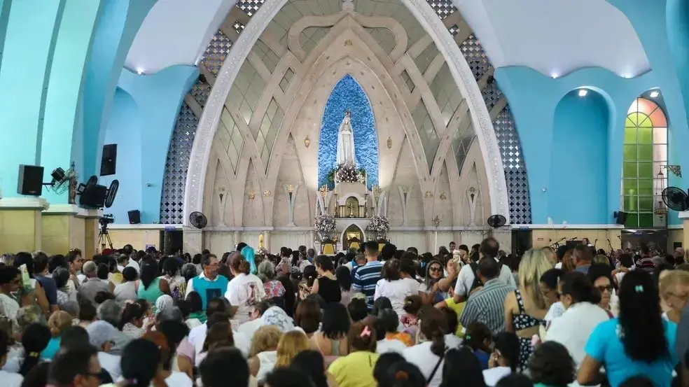 Na imagem, interior de uma igreja católica repleta de fiéis sentados e de pé durante uma celebração. Ao fundo, destaca-se o altar-mor emoldurado por grandes arcos ogivais entrelaçados em tons de bege e detalhes espelhados. No centro do altar, dentro de um nicho oval com fundo azul texturizado, está a estátua de Nossa Senhora de Fátima. As paredes laterais da igreja são pintadas em um tom vibrante de azul-turquesa, com colunas e arcos estruturais que sobem em direção ao teto. O público, visto de costas em primeiro plano, preenche todo o plano inferior da imagem.