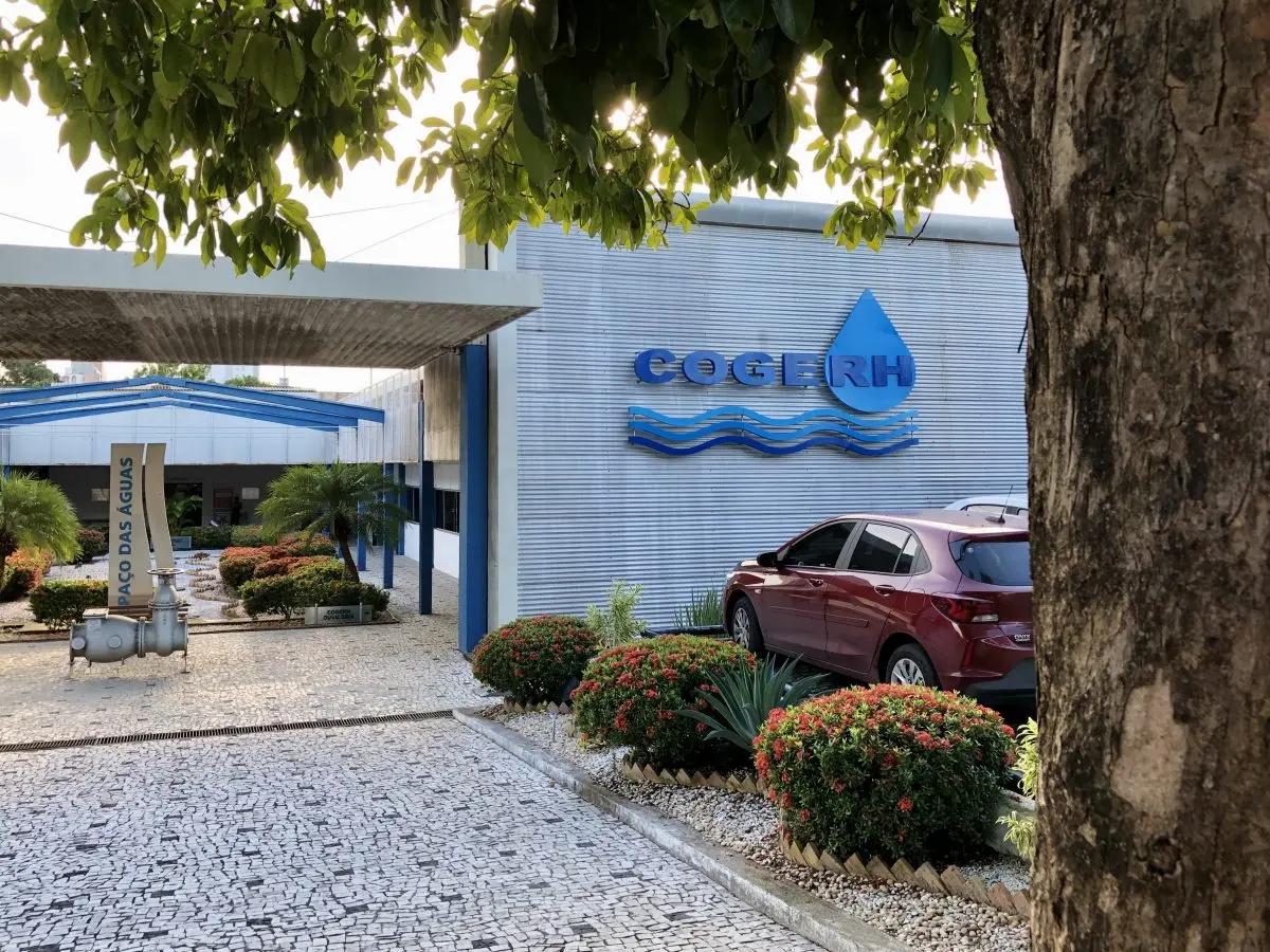 Vista da entrada da sede da Cogerh, com fachada metálica cinza exibindo o logotipo azul da instituição. Em frente ao prédio há um jardim com arbustos floridos e plantas ornamentais, além de um carro estacionado ao lado. À esquerda, uma estrutura coberta leva ao espaço identificado por uma placa com a inscrição “Paço das Águas”. A cena é enquadrada por uma árvore em primeiro plano e por folhas que cobrem parte superior da imagem.