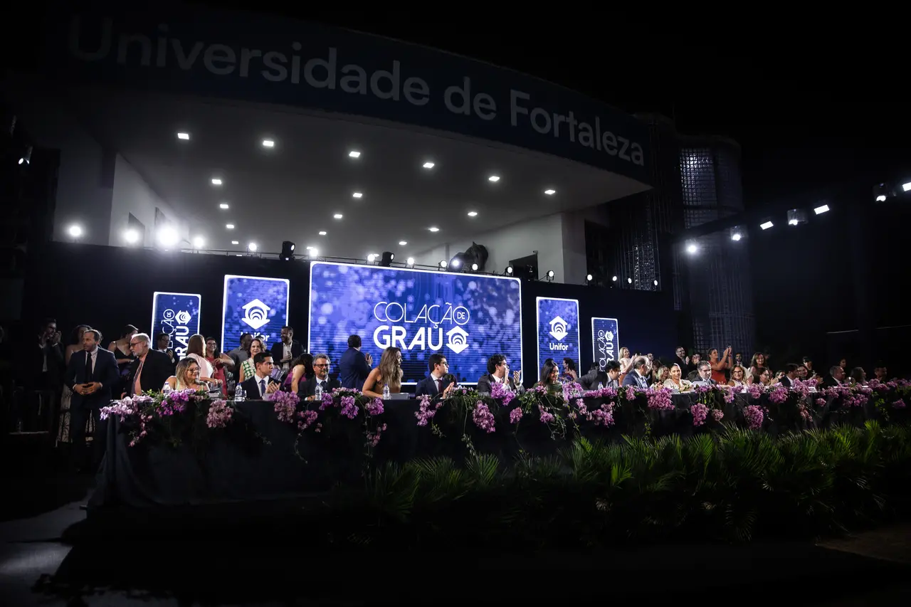 Uma longa mesa diretora adornada com flores roxas estende-se em um palco iluminado durante uma cerimônia de colação de grau noturna. Ao fundo, grandes painéis de LED azuis exibem o texto 'COLAÇÃO DE GRAU' sob uma estrutura da fachada que traz a inscrição 'Universidade de Fortaleza'.
