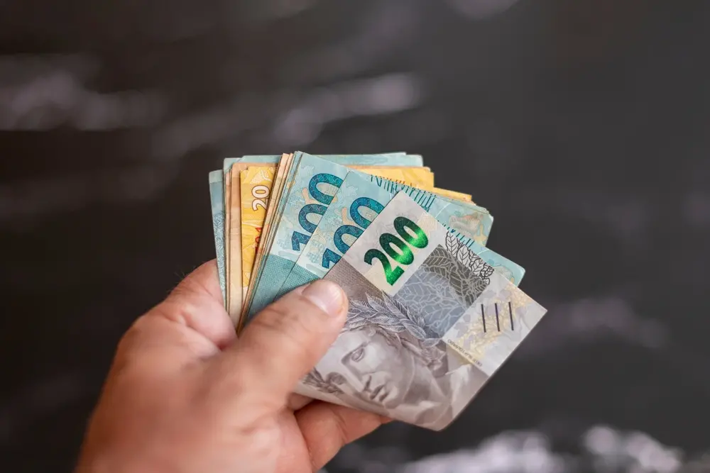 Mão segurando várias cédulas de real brasileiro, incluindo notas de R$ 200, R$ 100, R$ 20 e outros valores, com o dinheiro aberto em leque diante de um fundo escuro desfocado.