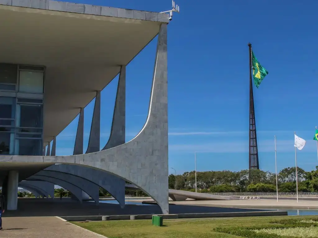 Fachada do Palácio do Planalto, com colunas de pedra e mastro à direita com bandeira do Brasil hasteada.