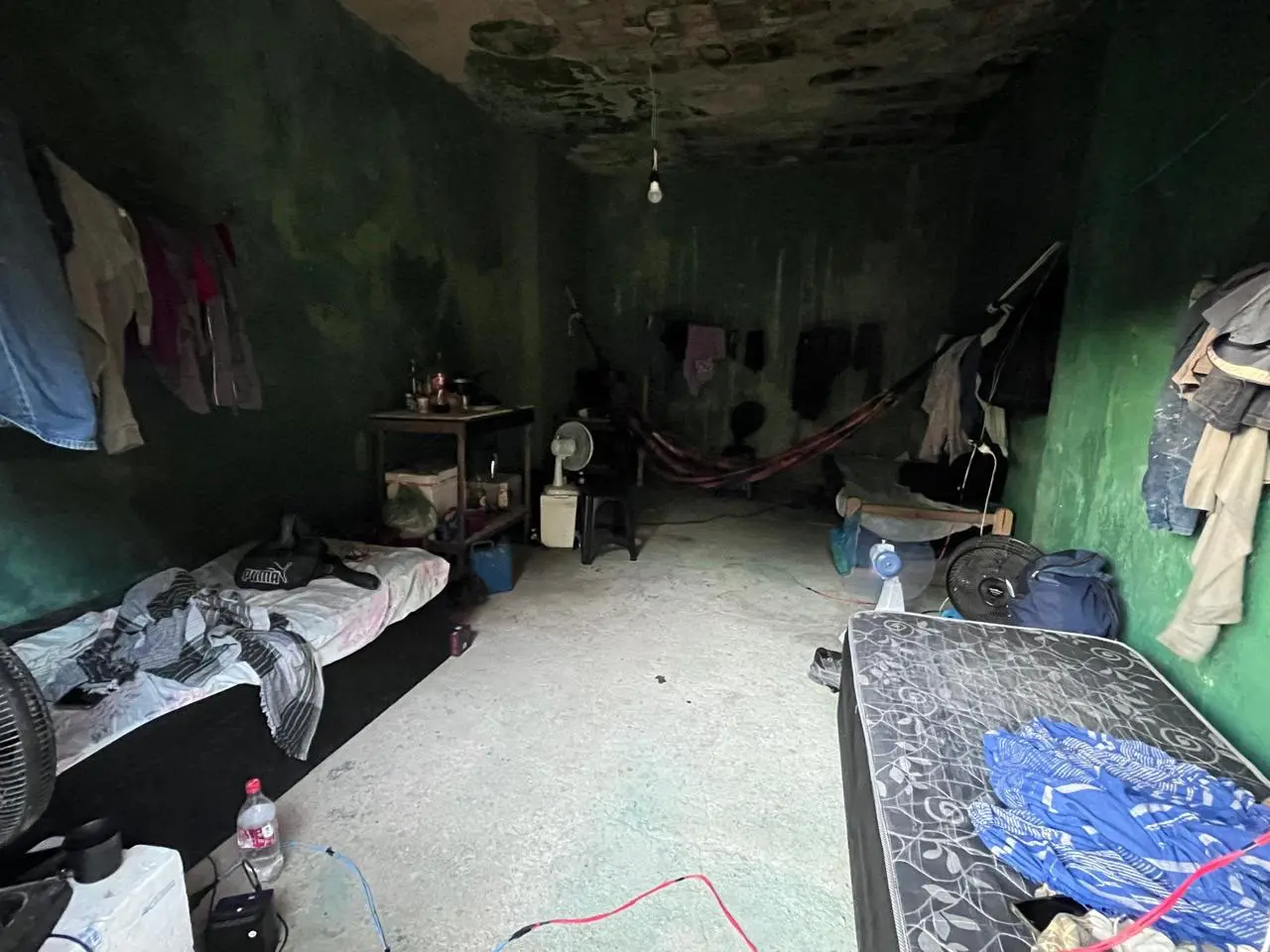 A imagem mostra interior de um alojamento precário e improvisado, com paredes verdes desgastadas, teto manchado e chão de cimento batido. No ambiente escuro e desorganizado, há colchões dispostos pelas laterais, roupas penduradas, ventiladores de chão, fiações expostas e uma rede armada ao fundo sob uma lâmpada fraca pendurada.