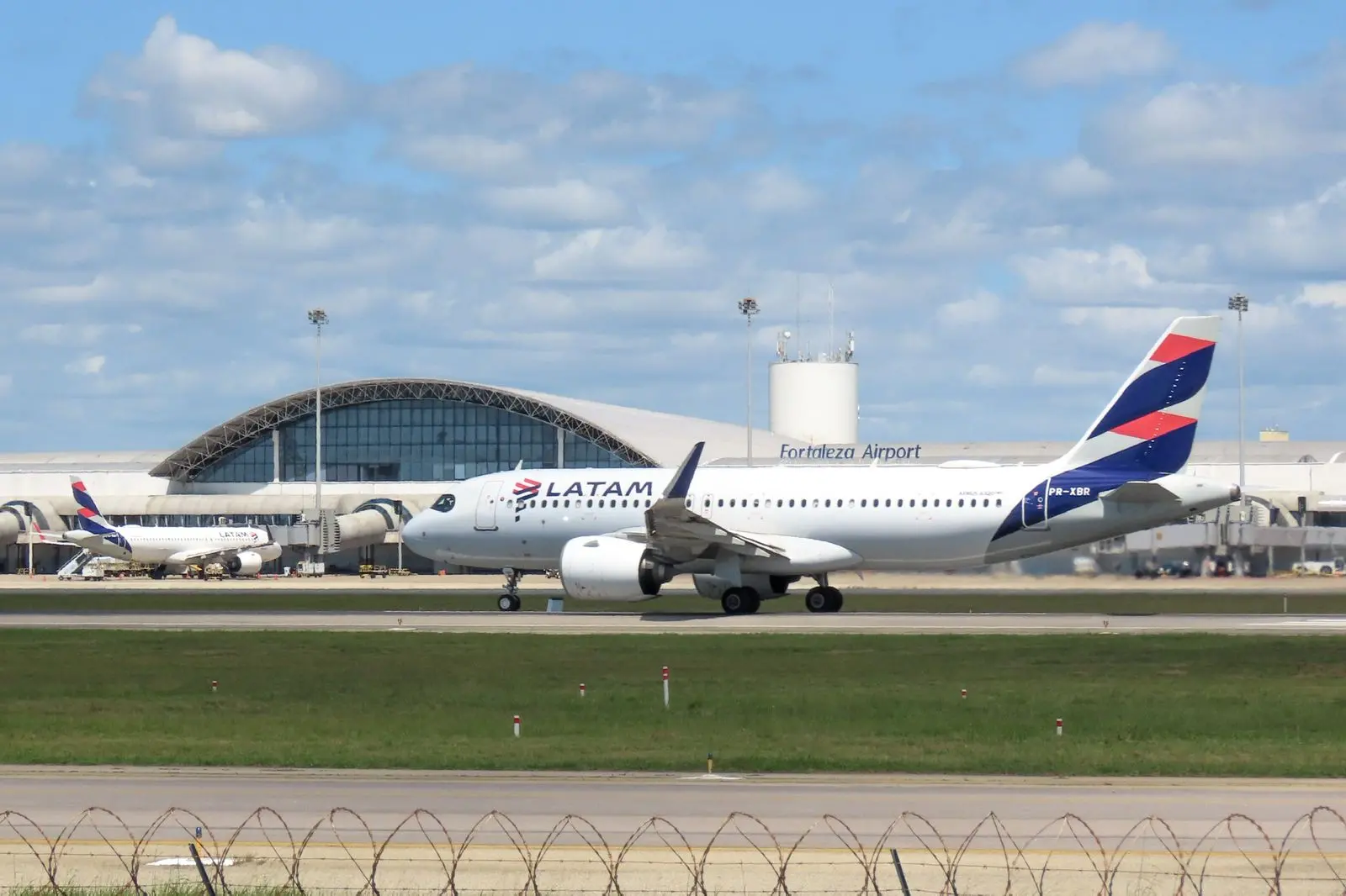 imagem mostra avião da latam pousando no aeroporto de fortaleza. Ao fundo, terminal do aeroporto.