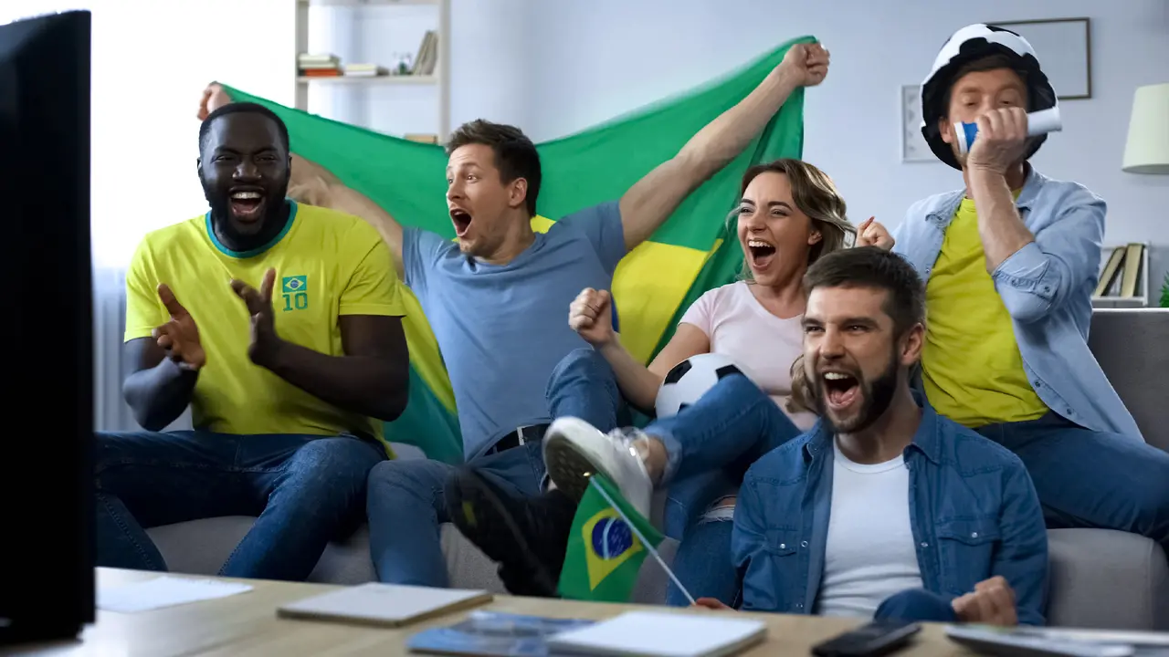 Um grupo de amigos felizes e descontraídos, numa sala decorada com as cores verde e amarelo, assiste ao jogo na televisão, rindo e partilhando petiscos.