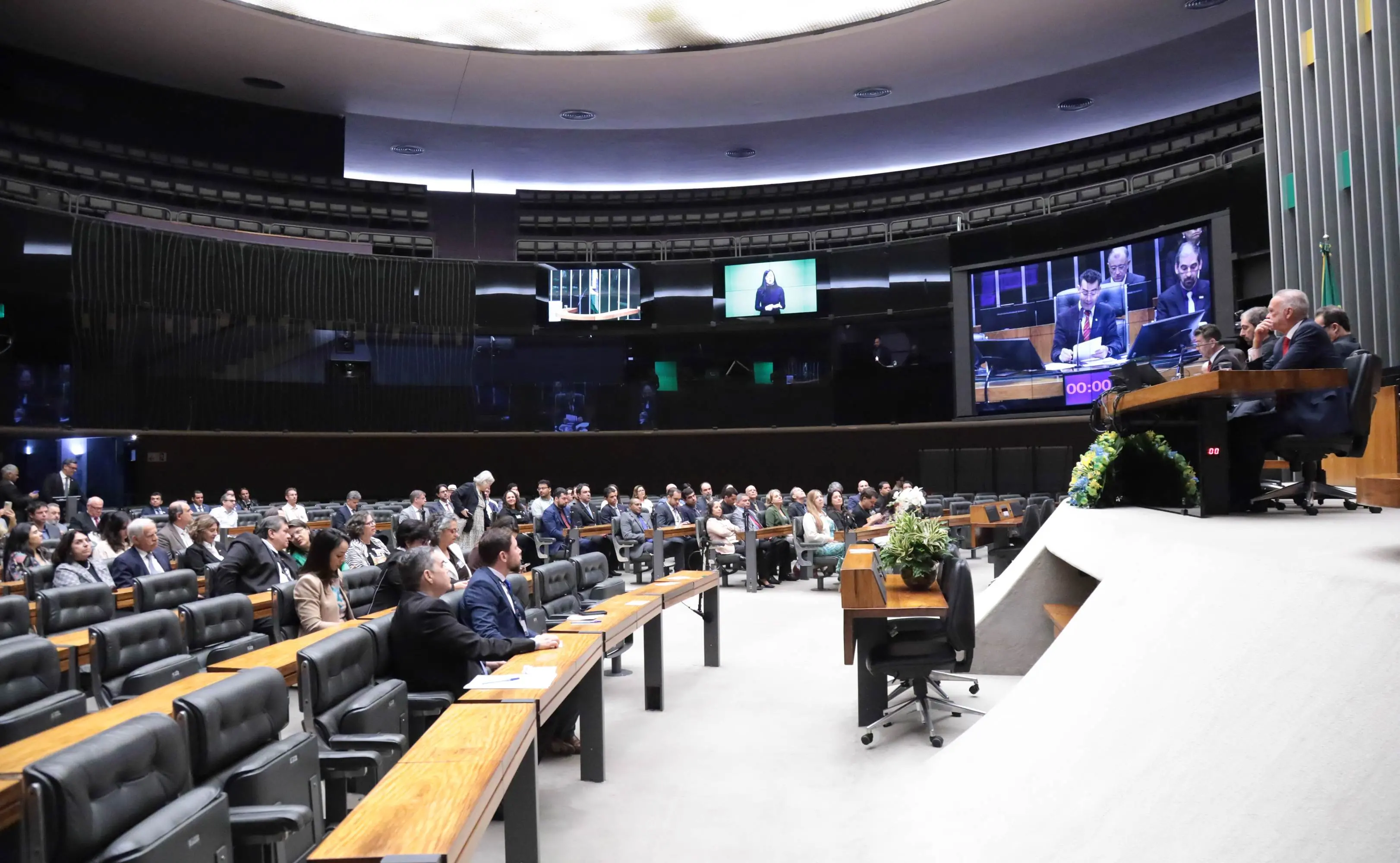 Plenário da Câmara dos Deputados durante sessão legislativa, com parlamentares e convidados presentes. À direita, integrantes da Mesa Diretora conduzem os trabalhos, enquanto o painel eletrônico exibe imagens da sessão.