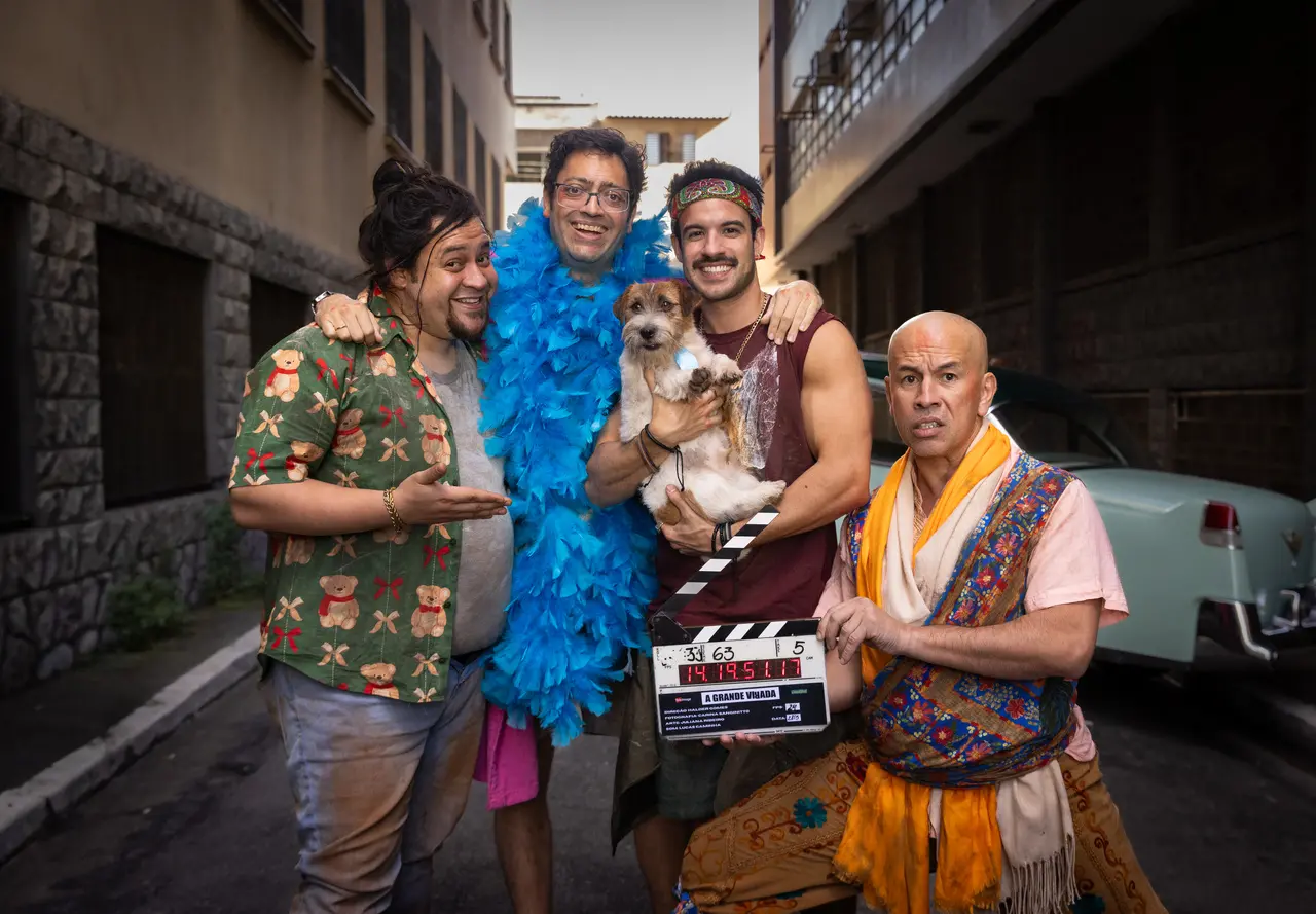 Cena de bastidores do filme A Grande Virada em uma rua estreita. Quatro pessoas fantasiadas e caracterizadas posam juntas diante de um carro antigo; uma delas segura um cachorro no colo e outra segura uma claquete de filmagem. Entre os figurinos, destacam-se uma estola azul de penas, roupas coloridas e adereços inspirados em diferentes estilos. Ao fundo, aparecem prédios e veículos estacionados.