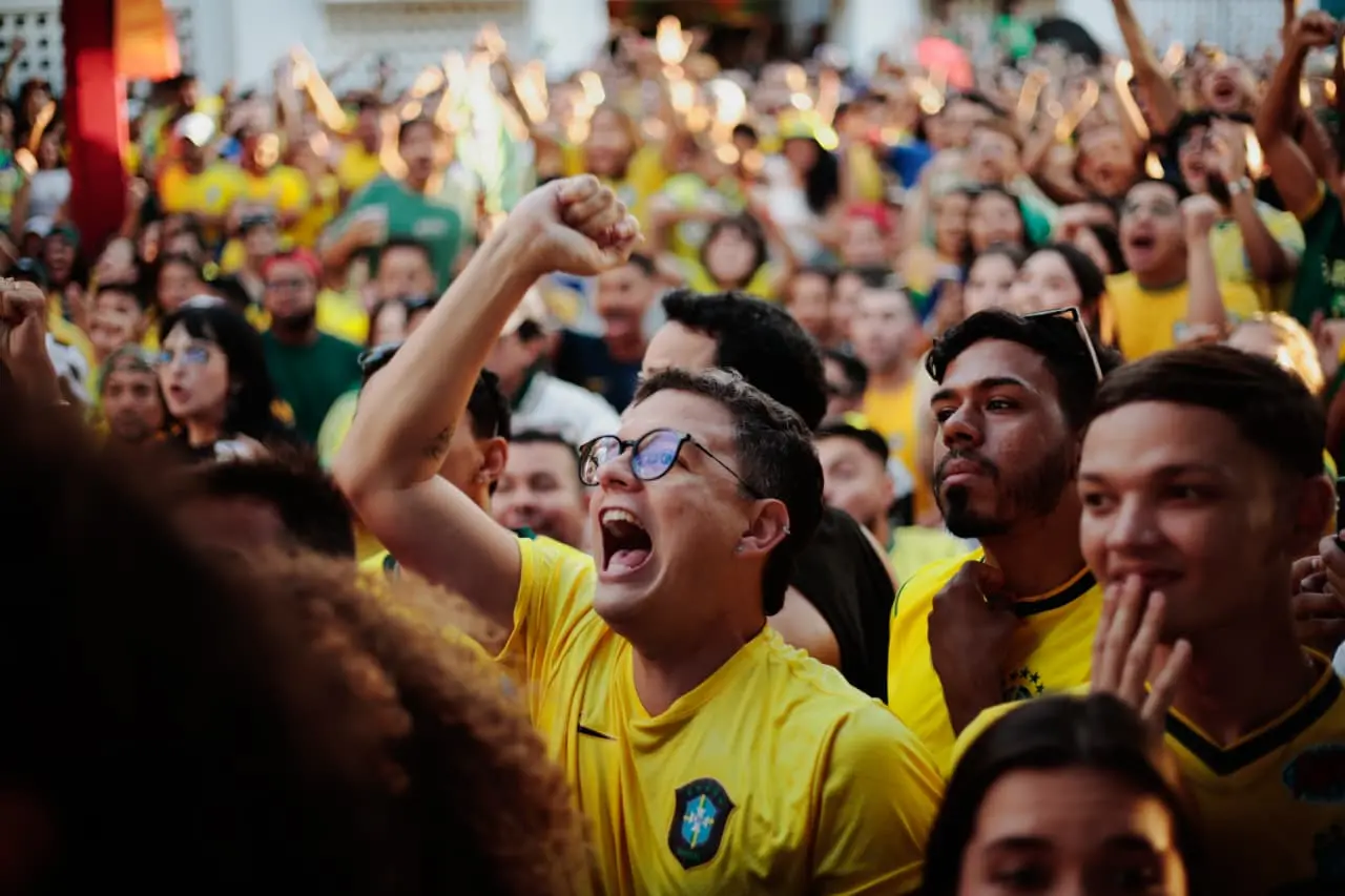 Um jovem de óculos e camisa amarela do Brasil grita e comemora com o punho erguido em meio a uma multidão de torcedores eufóricos.