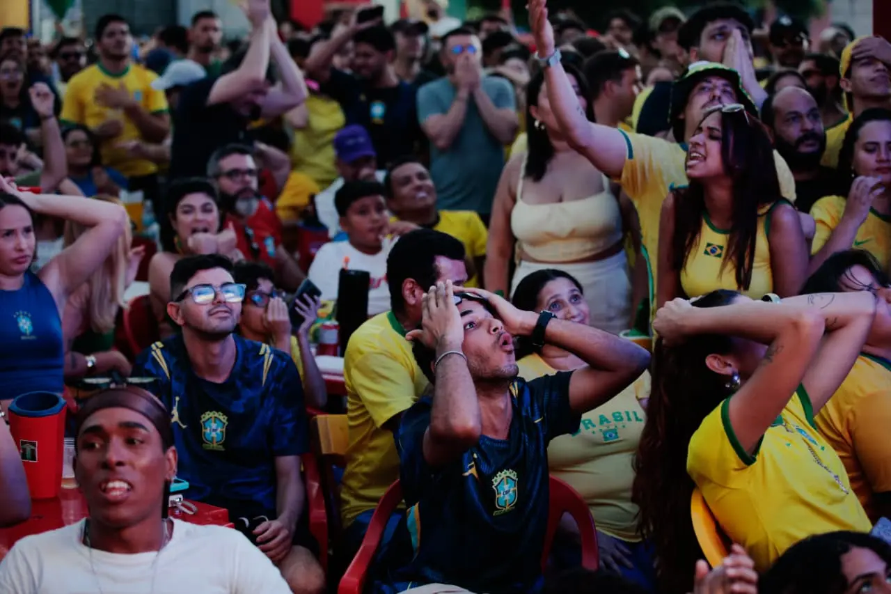 Torcedores com camisas do Brasil assistem a um jogo com expressões de forte tensão e apreensão; um homem ao centro leva as mãos à cabeça em desespero.
