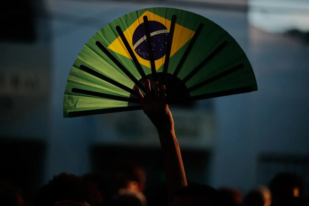 Uma mão erguida segura um leque aberto com a estampa da bandeira do Brasil, destacado contra um fundo escuro e desfocado no fim de tarde.