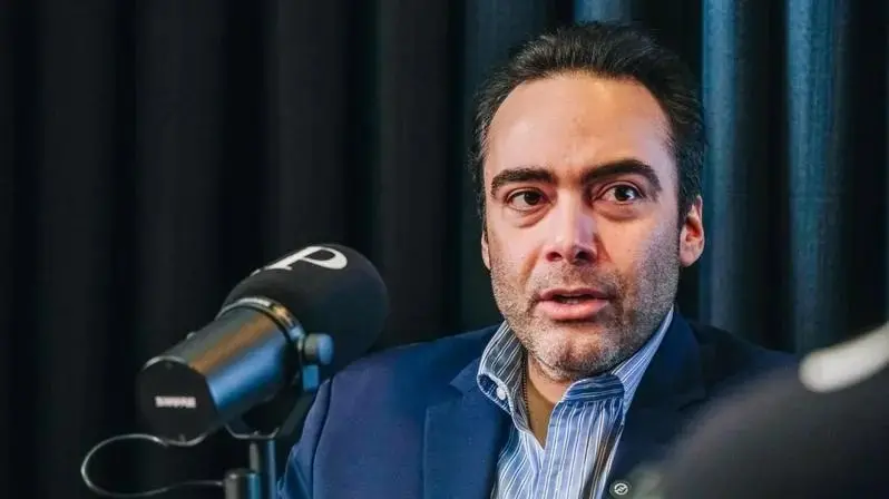 A imagem mostra um homem de terno azul e camisa social listrada em tons claros, falando diante de um microfone preto com a letra “P” em destaque na espuma protetora. Trata-se de Ozires Pontes. Ele tem pele clara, cabelos curtos e escuros, penteados para trás, além de barba cerrada. Ao fundo, há uma cortina escura desfocada, sugerindo que a cena ocorre em um estúdio de gravação ou entrevista.